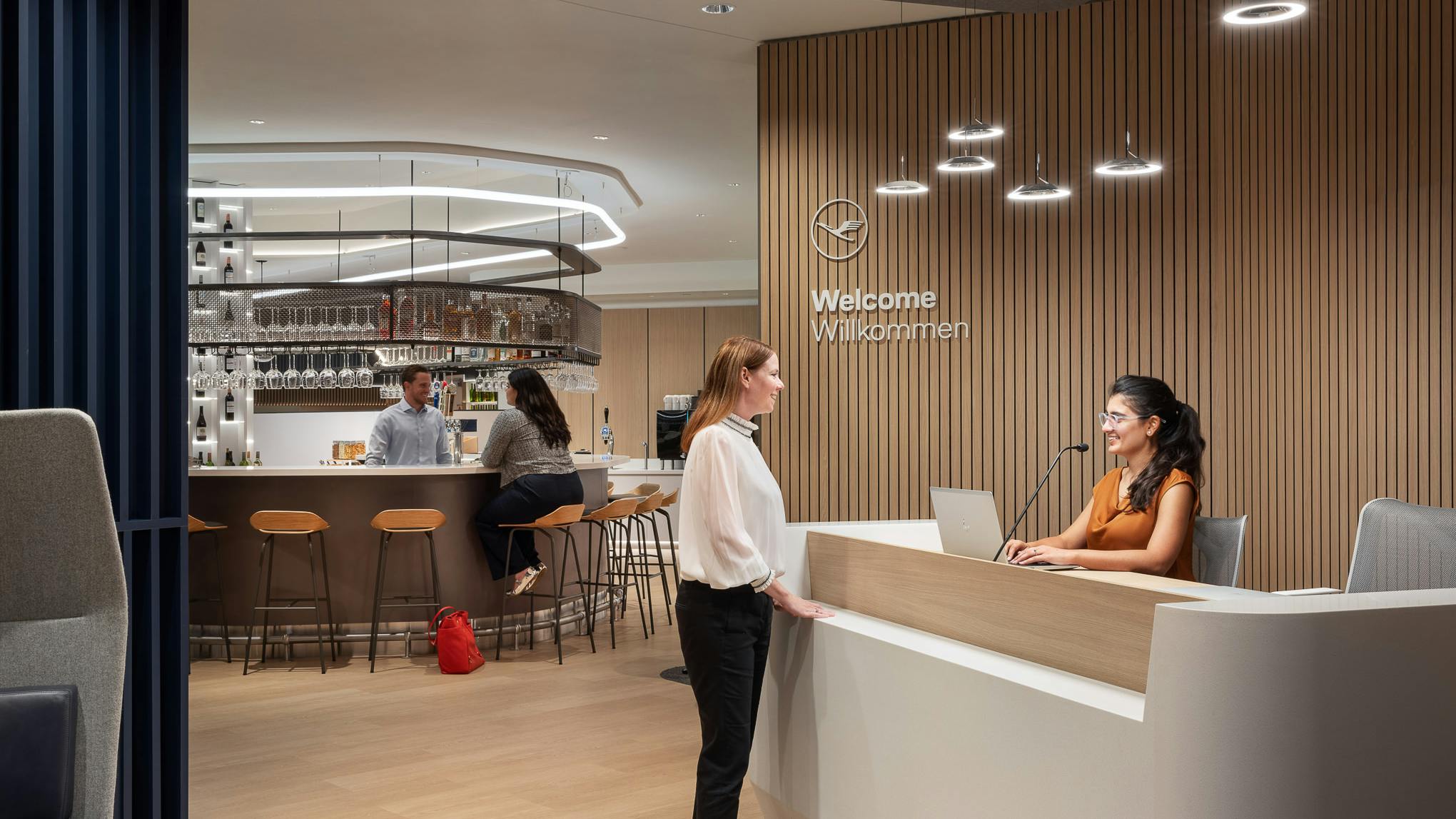 Woman standing at reception desk at the Lufthansa airlines lounge talking to receptionist with bar area in the background.