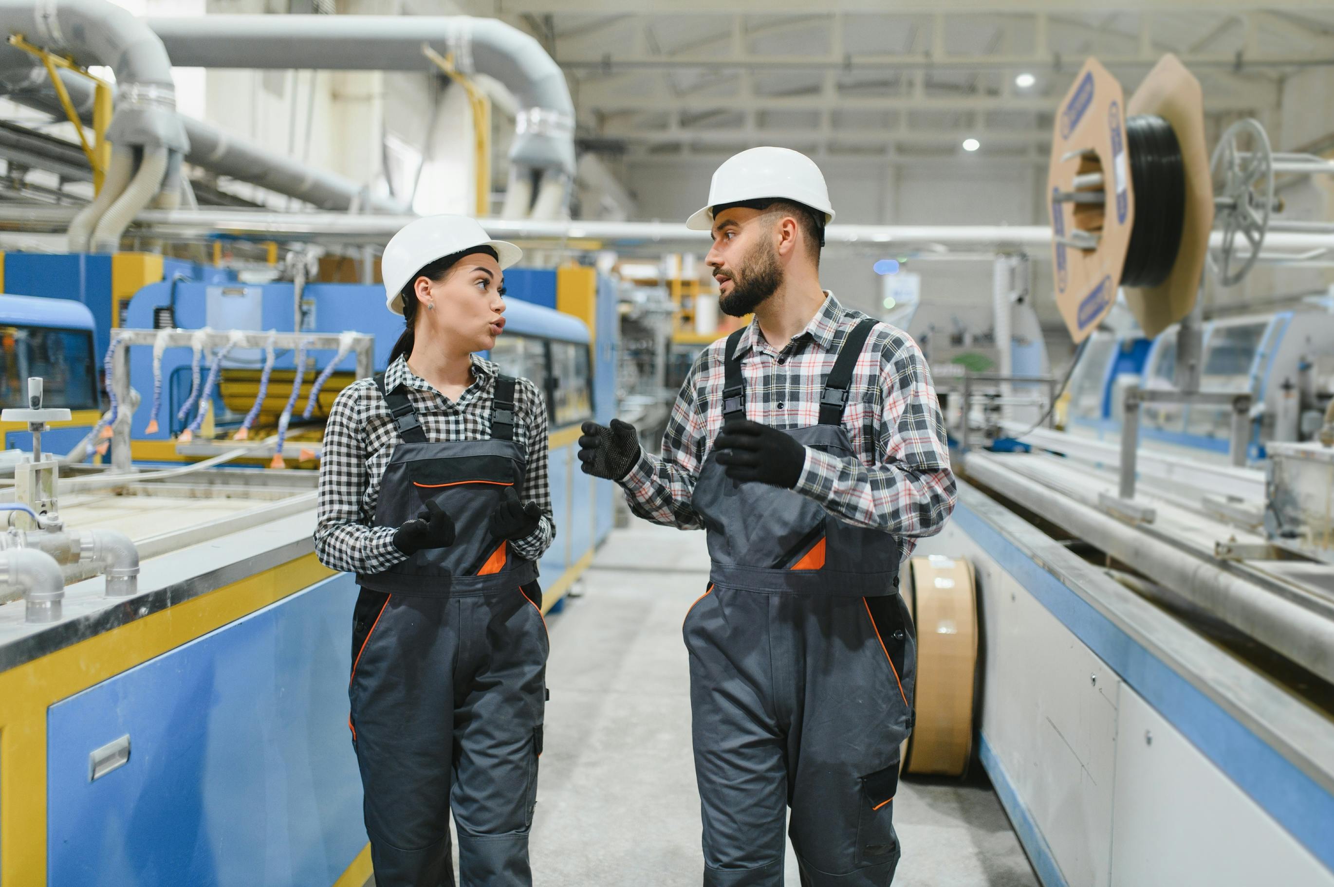 Two factory workers in overalls and hard hats are walking through a production line area.