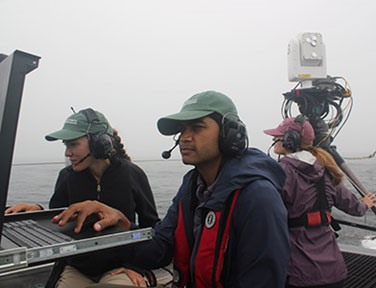 Johns Hopkins University Applied Physics Laboratory (APL) free-space optical (FSO) operators check the monitors on Sea Hunter as a third operator monitors operations. The FSO system can be seen at top right.