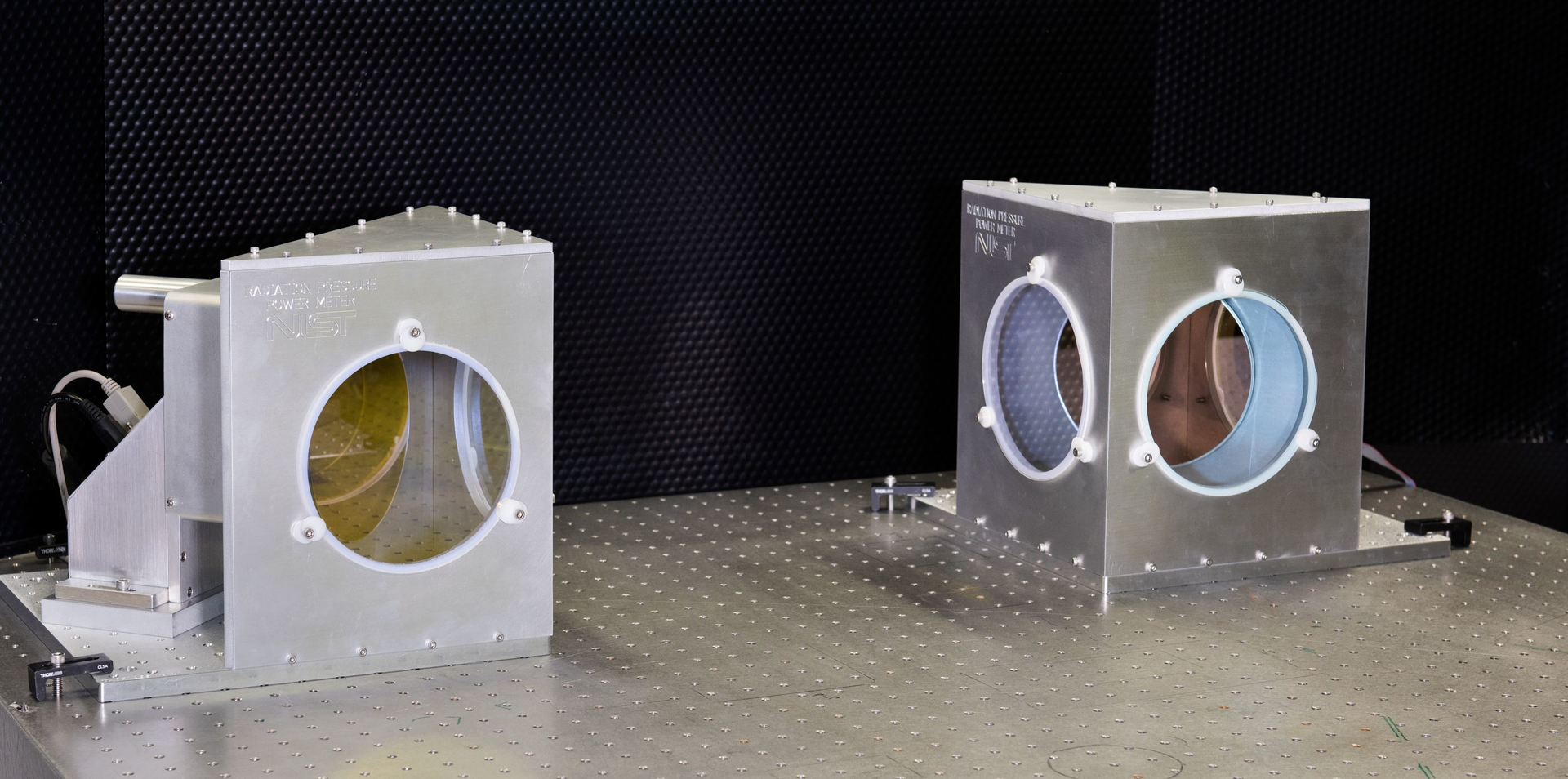 Each of these two NIST radiation pressure power meters (RPPMs) consists of a metal housing within which is a mirror connected to a vertical balance. From this angle, the mirrors appear slightly yellow (left) and pink (right).