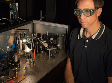 NIST engineer Tasshi Dennis sits next to NIST's solar simulator based on a white-light supercontinuum laser. The instrument simulates sunlight to help measure the properties of solar cell materials. The instrument's beam is illuminating a gallium arsenide solar cell in the lower left corner of the photo.