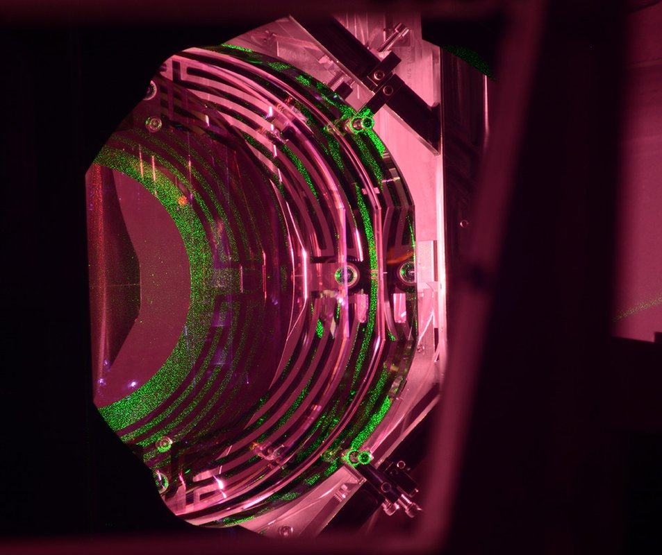 An Advanced LIGO optic has a mass of about 40 kg; about half of the cylindrical optic can be seen. The green light comes from an alignment laser. The maze-like patterns are actuators that translate, tip, and tilt the optic to maintain its alignment.