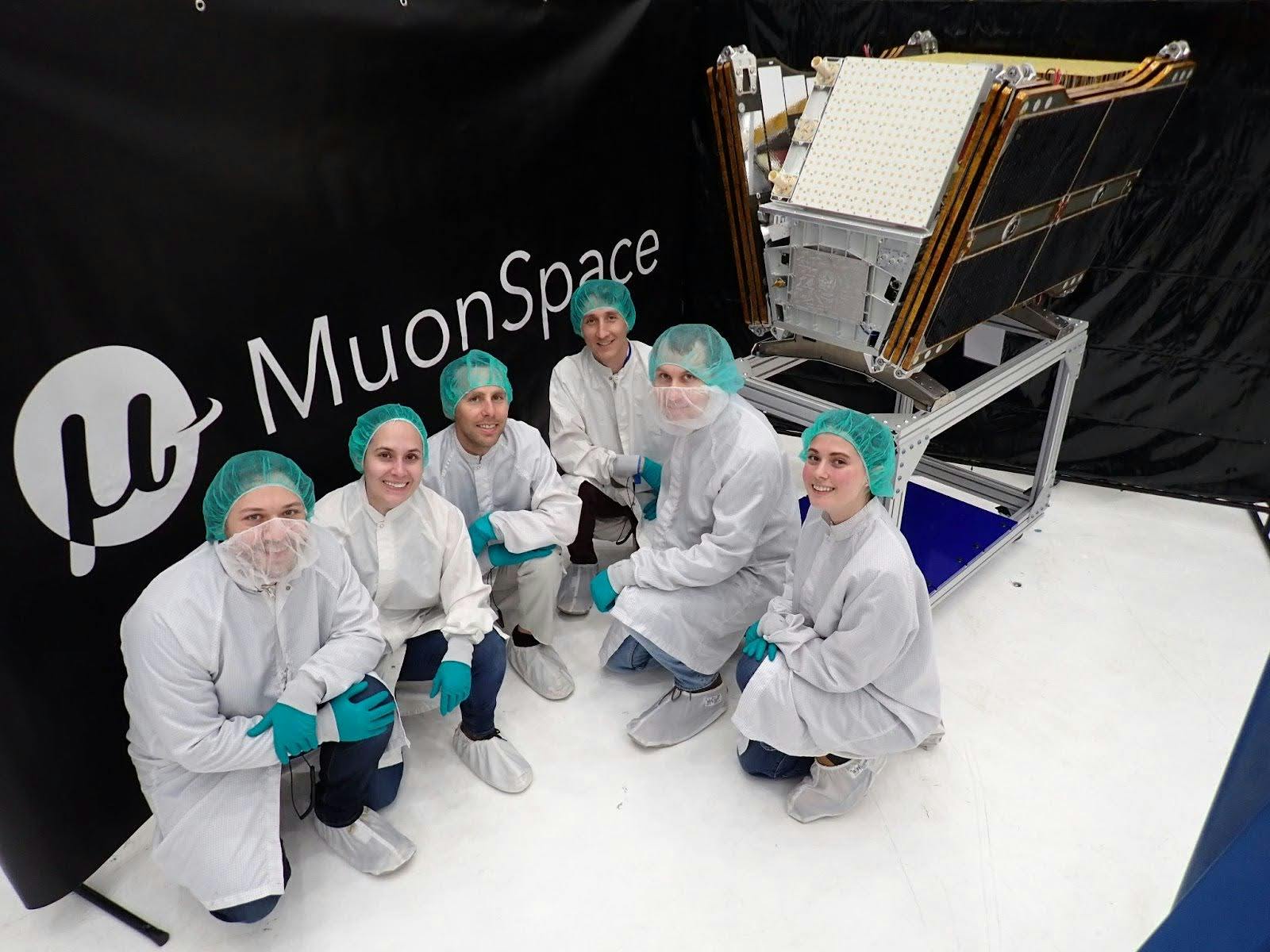 A group of people in cleanroom clothing pose in front of a satellite