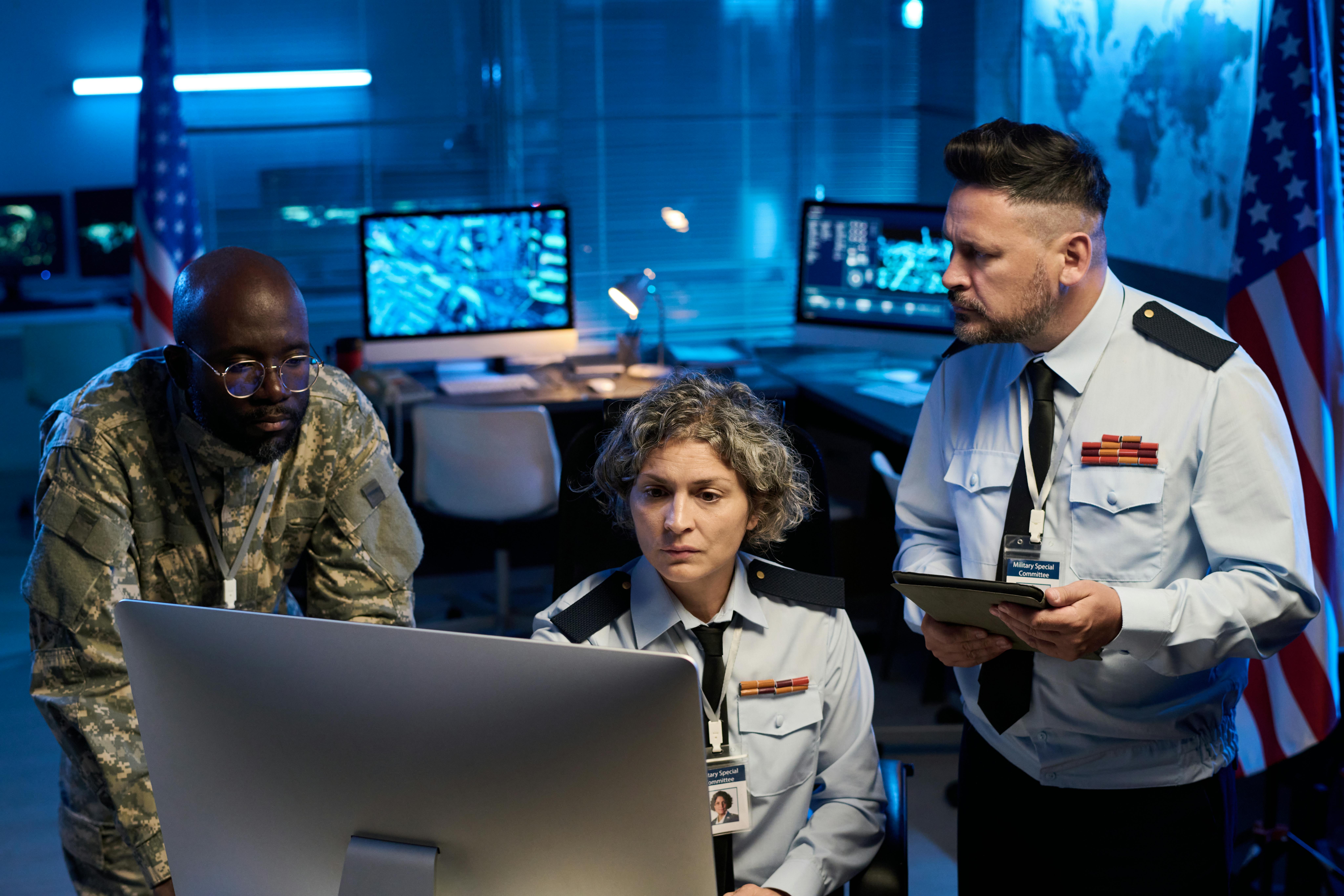 Team of serious workers of military command center looking at online data while sitting and standing in front of computer monitor