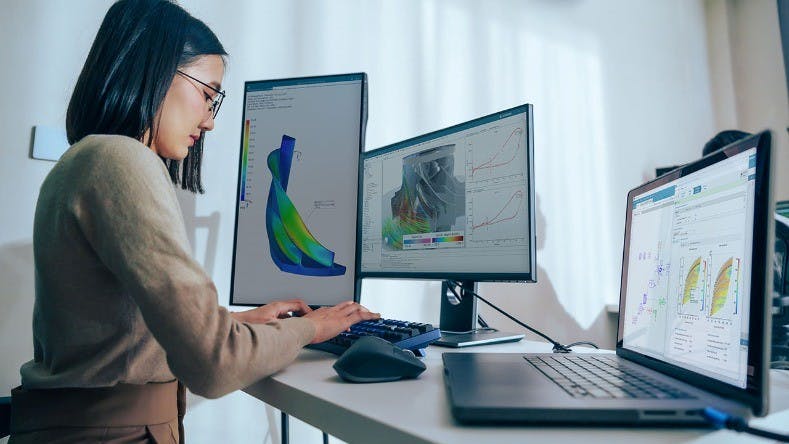 A woman works at a desk on a computer keyboard with a laptop and two monitors in front of her