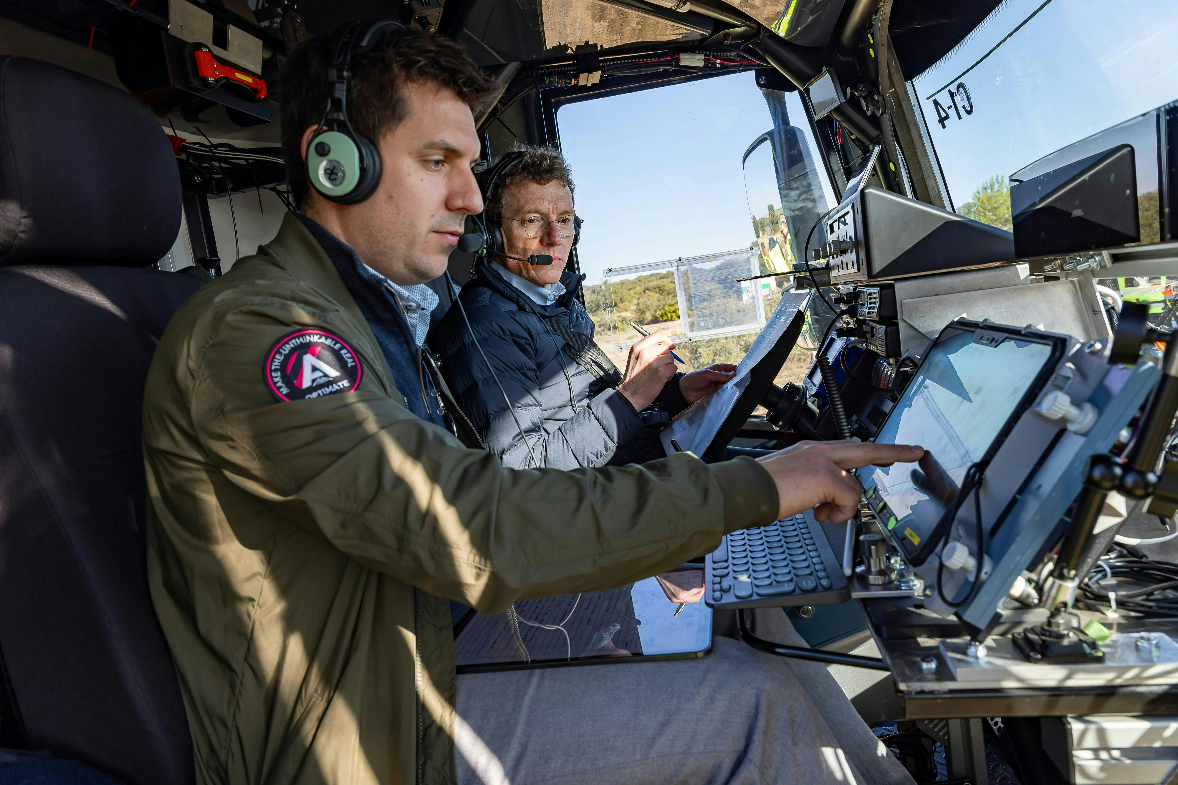 A man working on a tablet inside a vehicle while another looks on
