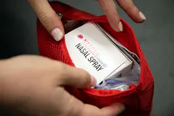Close-up of a person's hands holding a red bag with a box of naloxone nasal spray inside. National Institute on Drug Abuse. Close-up of a person's hands holding a red bag with a box of naloxone nasal spray inside. National Institute on Drug Abuse.