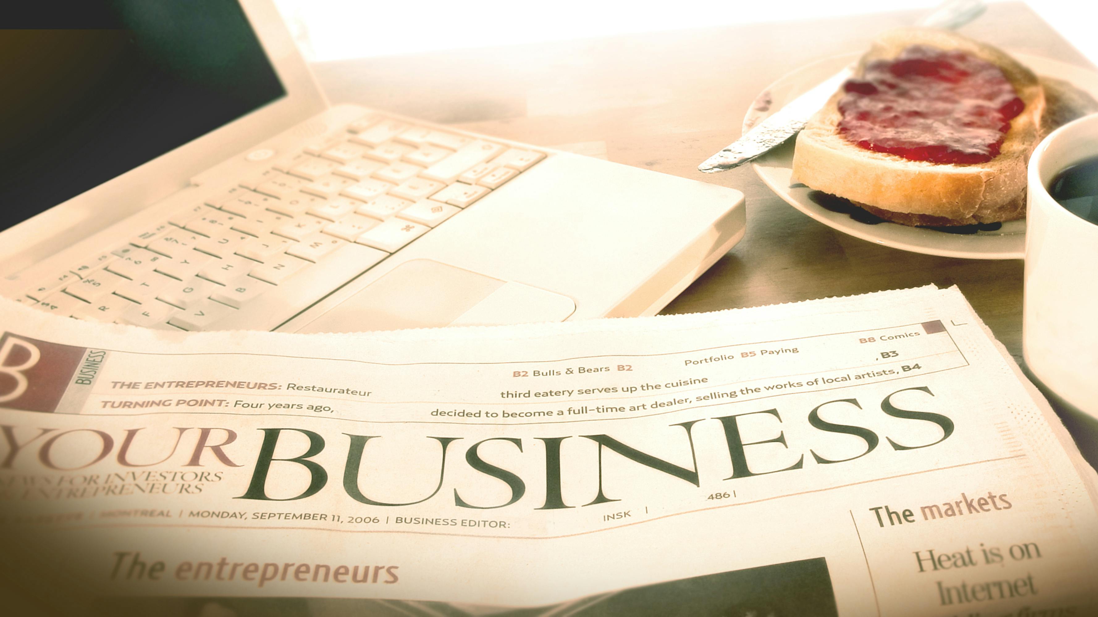 Newspaper business section sitting at desk with laptop and breakfast.