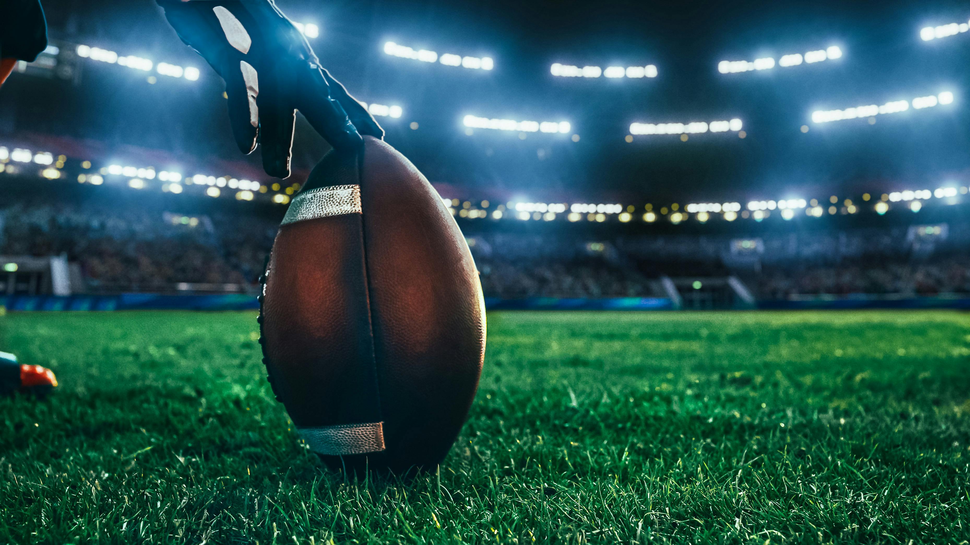 close-up shot of an American football being held by a football player on the stadium field at night with lights and crowd in the background.