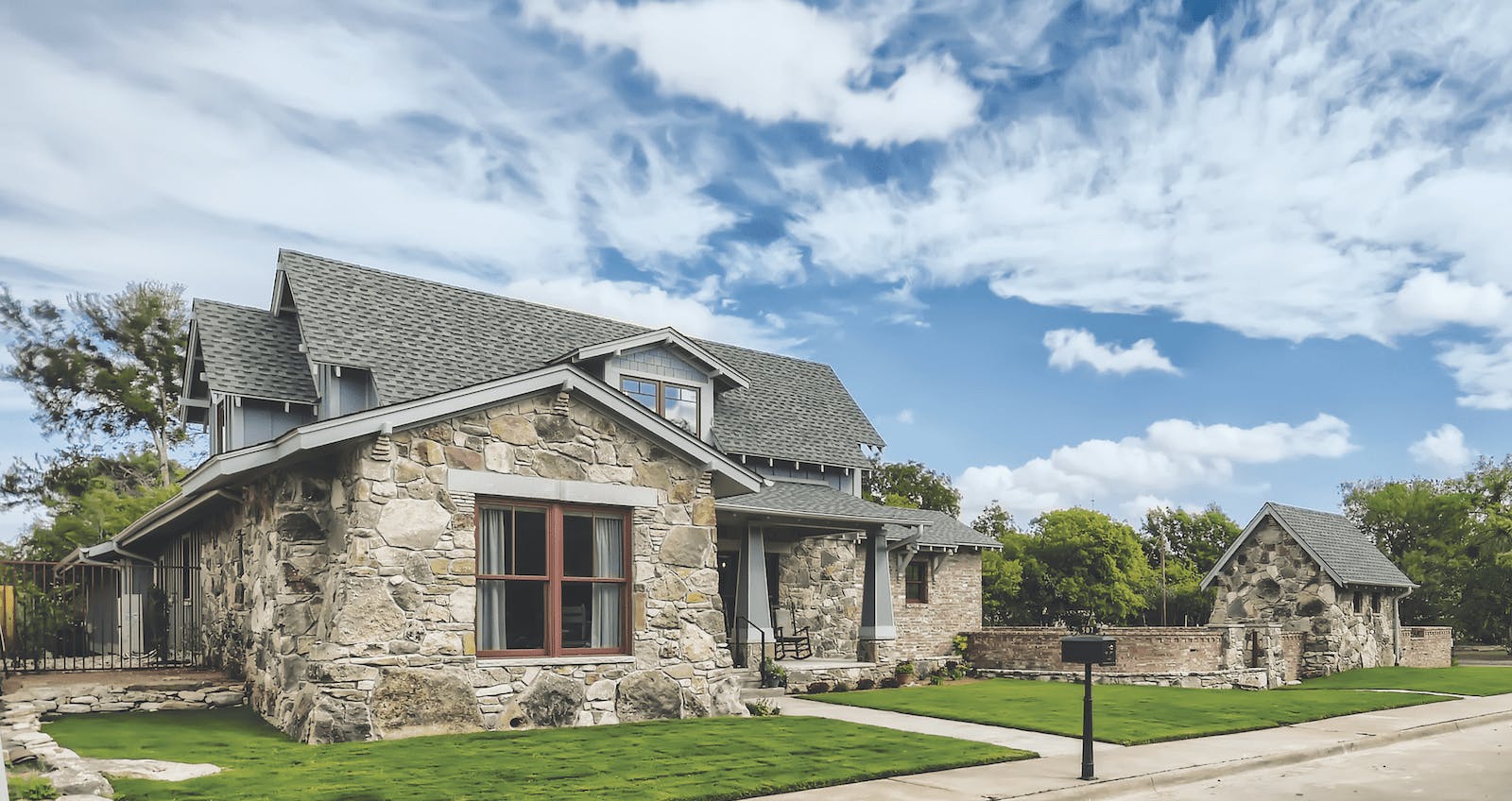 The extra-wide street frontage allowed space for a detached Studio, while a gently sloping walkway to the home&rsquo;s raised front porch offers an &ldquo;access-friendly&rdquo; entrance. Stone window headers, low-profile gutters, and slivers of tapered stone just below the roofline are small but impactful details in this Arts and Crafts&ndash;inspired design.