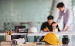 A student and teacher go over an assignment in the background. In the foreground, there is a table with some papers and a hard hat. A student and teacher go over an assignment in the background. In the foreground, there is a table with some papers and a hard hat.