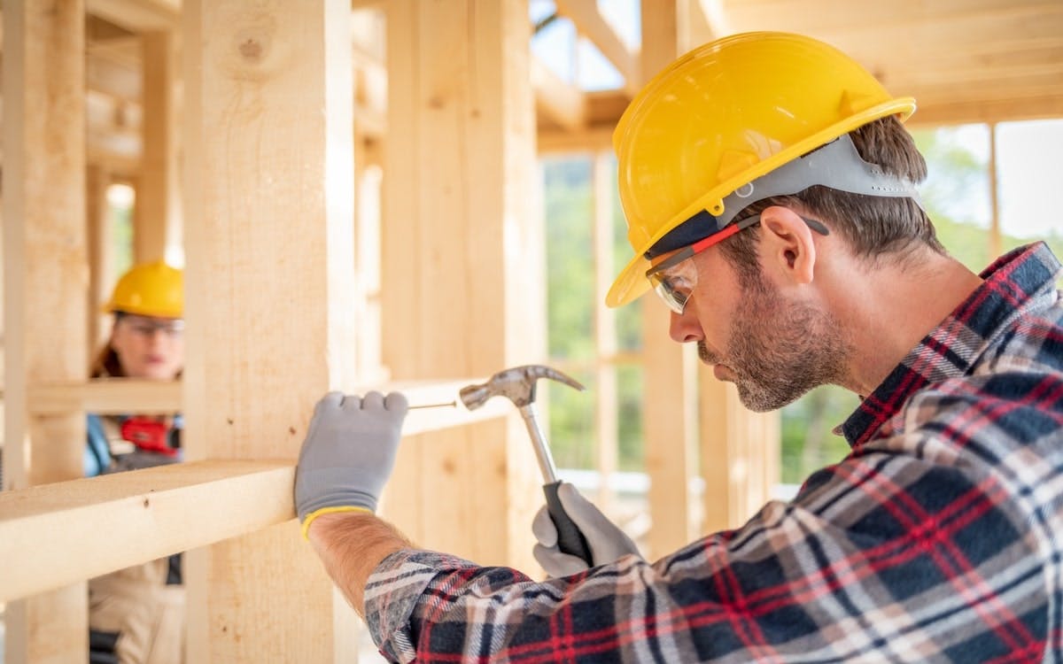 A man in a yellow hard hat hammers a nail into the framing of a new home