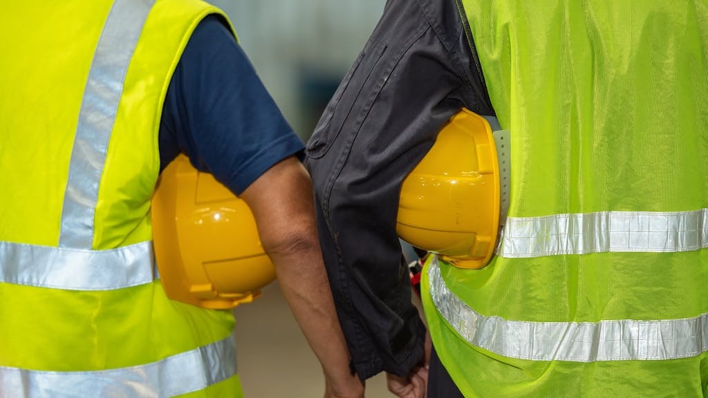 Two construction workers wearing reflective vests and carrying hard hats