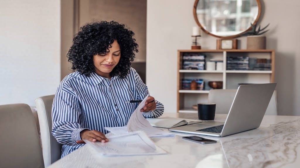 A remote worker looks over notes with her laptop open on the table in front of her.