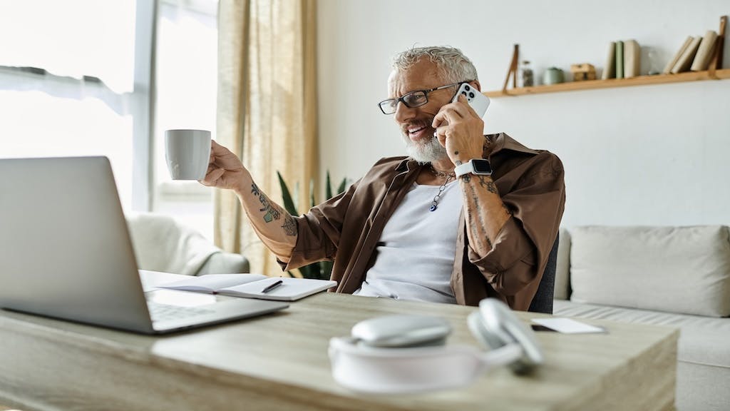 A Gen-X man talks on the phone while looking at a laptop and drinking coffee.