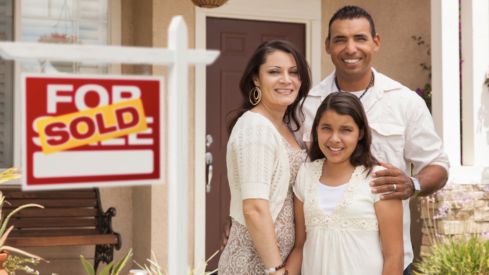 A couple with their daughter stand in front of their home with a 'sold' sign
