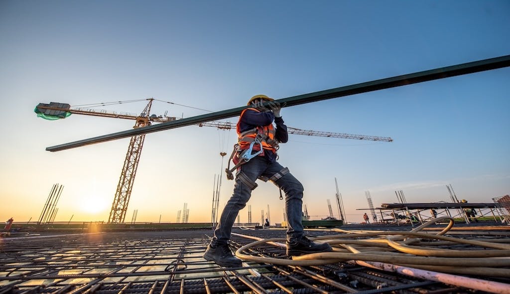 A construction worker walks over rebar carrying a beam on his shoulder.