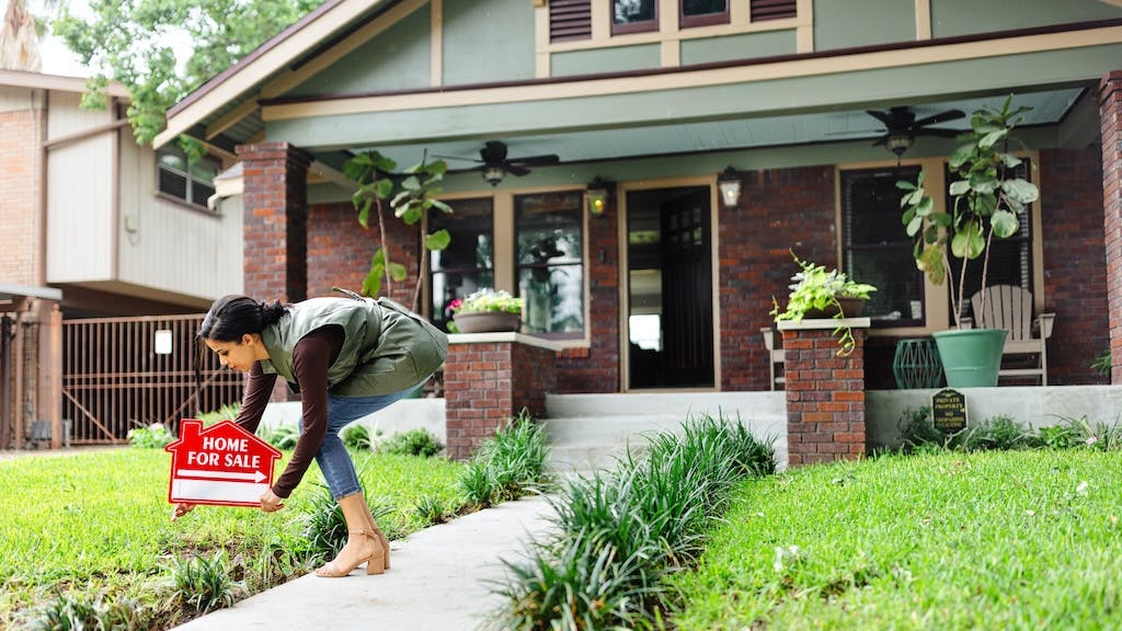 A woman places a 'For Sale' sign in the front yard of a home