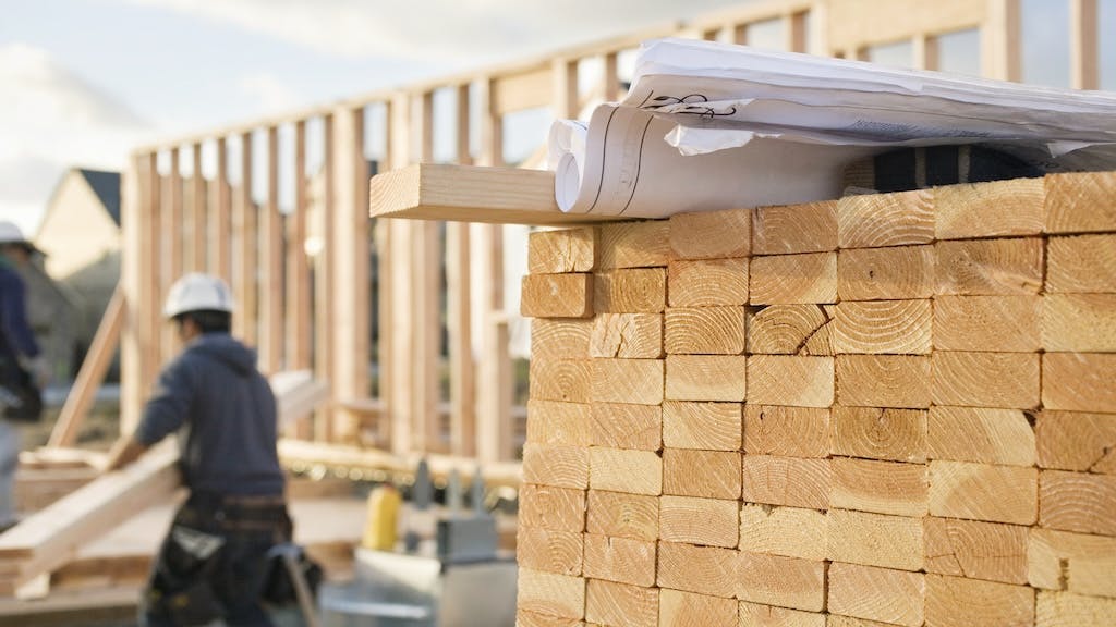A stack of lumber with a builder working on a home in the background