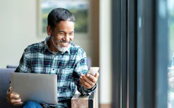 A Man In A Plaid Shirt Smiles While Holding His Laptop In One Hand And Looking At His Phone In The Other A Man In A Plaid Shirt Smiles While Holding His Laptop In One Hand And Looking At His Phone In The Other