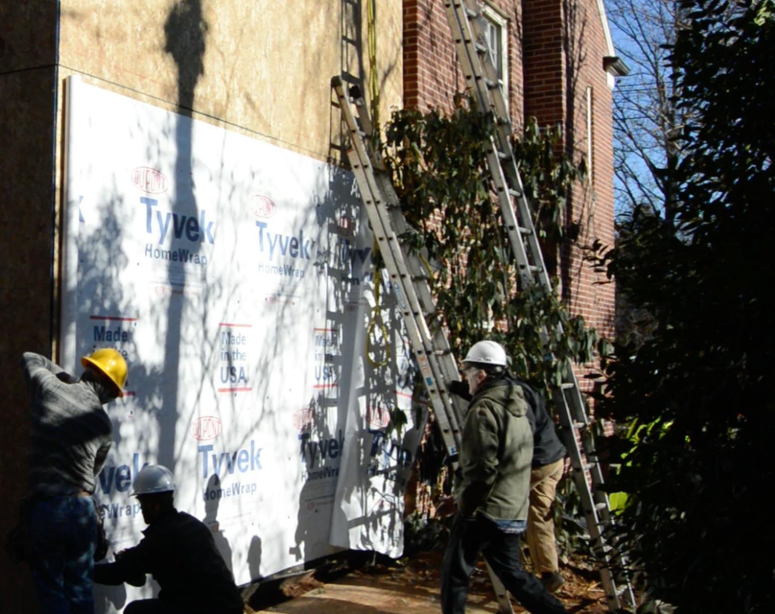 House wrap installation begins at the bottom. Tall rolls make smoothing the membrane easier, but they become unwieldy when climbing ladders. Upper courses are cut in advance and stretched corner to corner before nailing or stapling off.