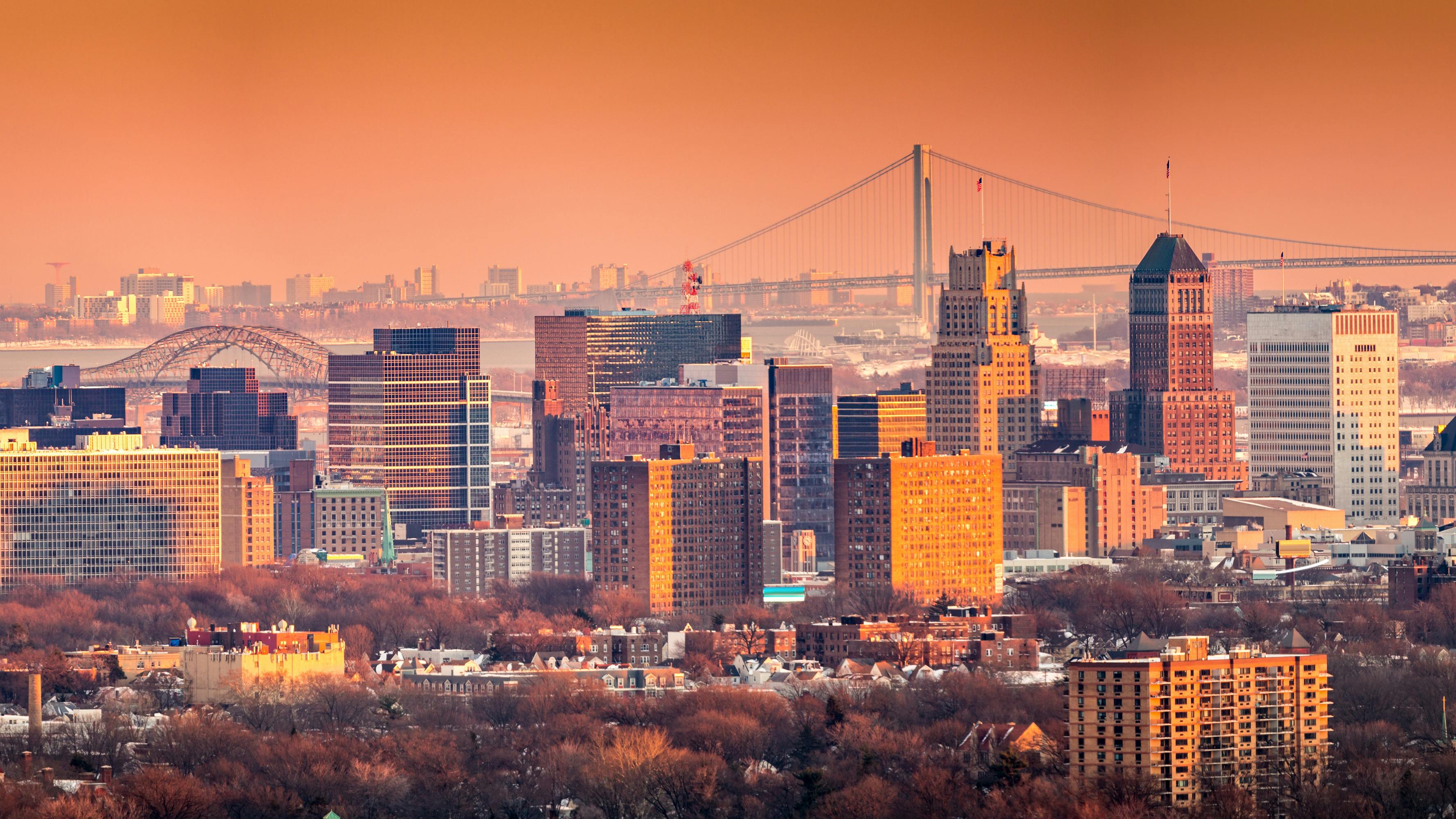 Newark New Jersey skyline viewed from Eagle Rock reservation under an orange sunset. In the background, under a hazy sky, Verrazano bridge links Staten Island to Brooklyn.